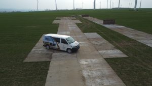 Biały furgon stoi na betonowej nawierzchni na zielonym polu, w tle widać wiatraki.
