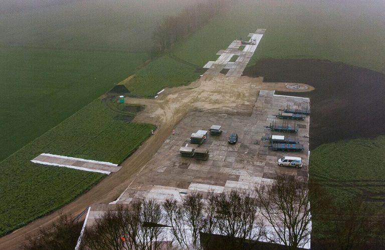 Widok z góry na plac budowy z płaską nawierzchnią, otoczony zielonymi polami i mgłą. W tle widoczne pojazdy i materiały budow