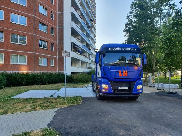 Niebieski ciężarówka z napisem LH parkuje przy budynku mieszkalnym, obok przystanku autobusowego.