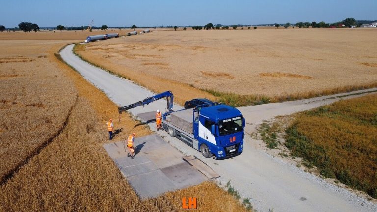 Na zdjęciu widać dużą ciężarówkę na wiejskiej drodze, obok niej pracują dwie osoby w odzieży roboczej. W tle pola.