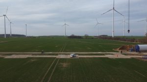 Widok na zielone pole z turbinami wiatrowymi w tle oraz samochodem na betonowej nawierzchni.