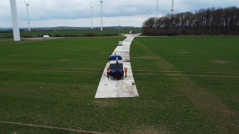 Na obrazie widać długi betonowy pas w zielonym polu, obok stoją samochody i pracownicy w pomarańczowych strojach.