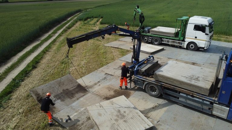 Na obrazku widać ciężarówkę z platformą, robotników w pomarańczowych strojach i dźwig przenoszący materiały. W tle pola.