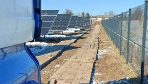 Widok na farmę paneli słonecznych wzdłuż wyłożonej drewnem ścieżki, z ogrodzeniem na końcu ścieżki.
