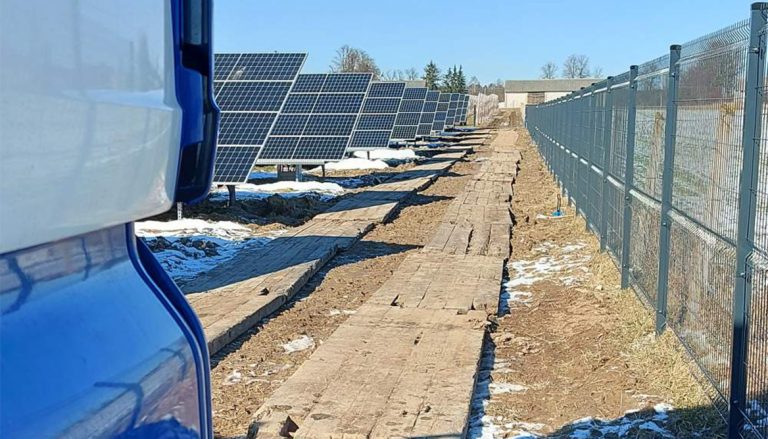 Widok na farmę paneli słonecznych wzdłuż wyłożonej drewnem ścieżki, z ogrodzeniem na końcu ścieżki.