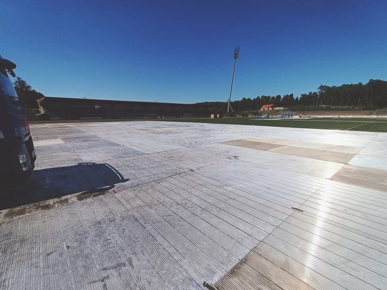 Pusta powierzchnia boiska pokryta panelami, otoczona zielenią i jasnym niebem. W tle widać oświetlenie stadionu.