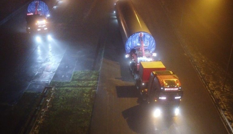Ciężarówki transportujące długie elementy w gęstej mgle, oświetlone reflektorami na drodze.
