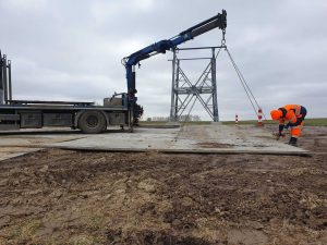 Pracownik w pomarańczowej odzieży roboczej przygotowuje teren budowy pod maszt, z dźwigiem i platformą.