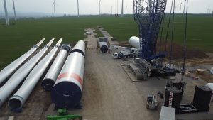 Na zdjęciu widać plac budowy turbin wiatrowych, z poziomymi wirnikami, ciężkim sprzętem i wiatrowymi elektrowniani w tle.