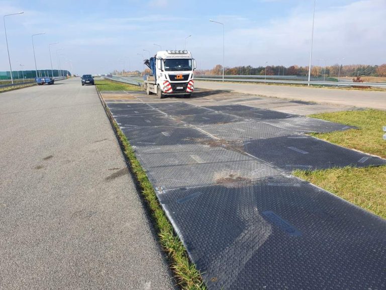 Ciężarówka na placu budowy obok drogi, na którym leżą płyty stalowe. W tle widoczna asfaltowa jezdnia.