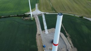 Widok na pole z wiatrakiem w budowie, betonową drogą i zielonymi uprawami w tle.