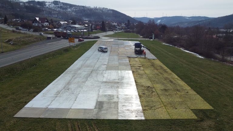 Widać plac budowy z częścią utwardzoną i pojazdami na zielonej trawie, w tle wiejskie osiedle i góry.