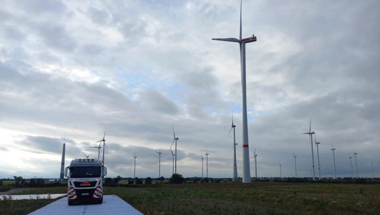 Ciężarówka na drodze w parku wiatrowym z wieloma turbinami w tle i pochmurnym niebem.