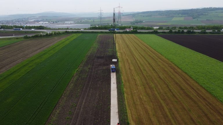 Widok na pola uprawne z widoczną drogą, ciągnikiem oraz różnorodnymi kolorami roślinności.