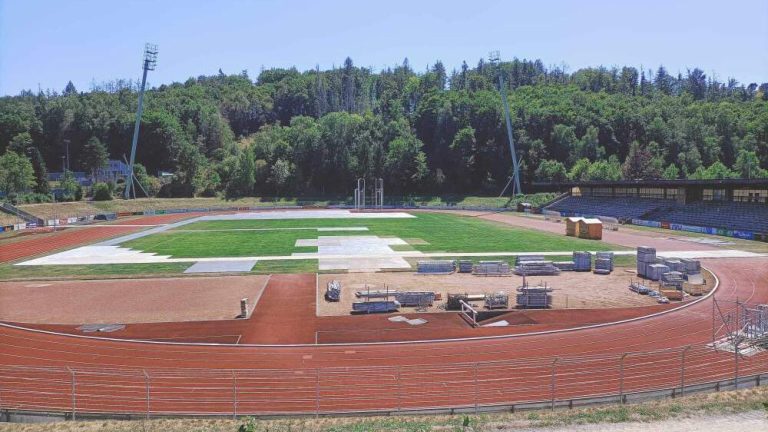 Stadion lekkoatletyczny z bieżnią, boisko w trakcie prac budowlanych, otoczony drzewami.