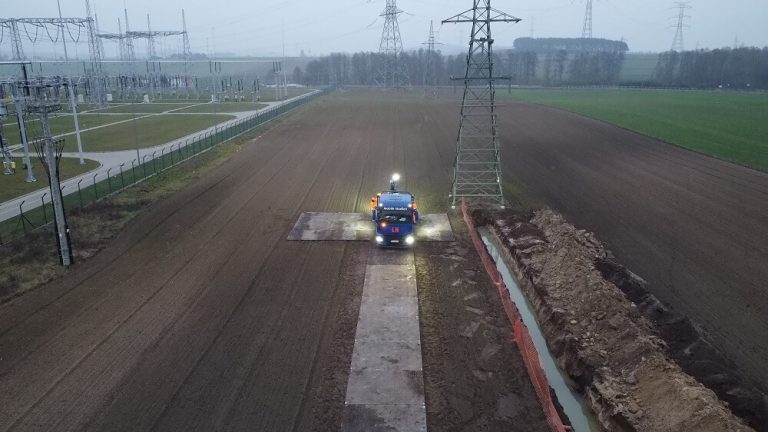 Widzimy teren w budowie z pojazdem budowlanym, pod liniami energetycznymi oraz z widocznym polem w tle.