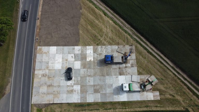 Widok z góry na plac utwardzony betonowymi płytami, z dwoma ciężarówkami i jednym samochodem, obok drogi.