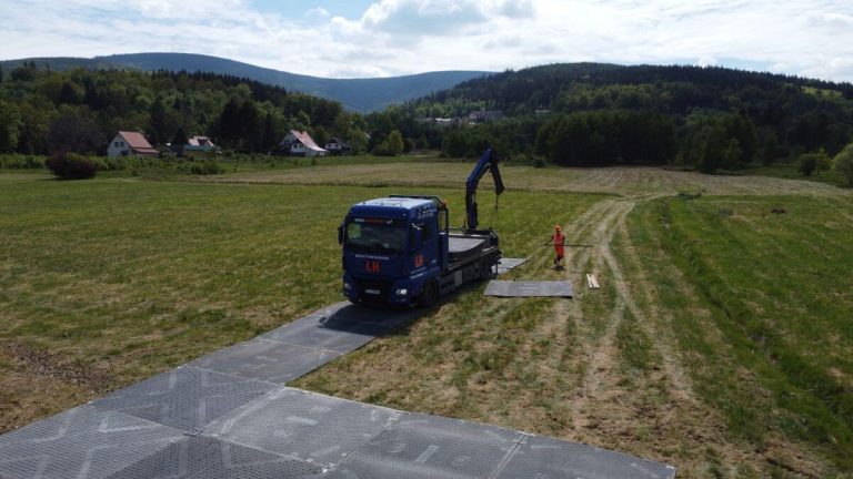 Na zdjęciu widoczny jest niebieski ciężarówka z dźwigiem na placu budowy w zielonym terenie, w tle lasy i domy.