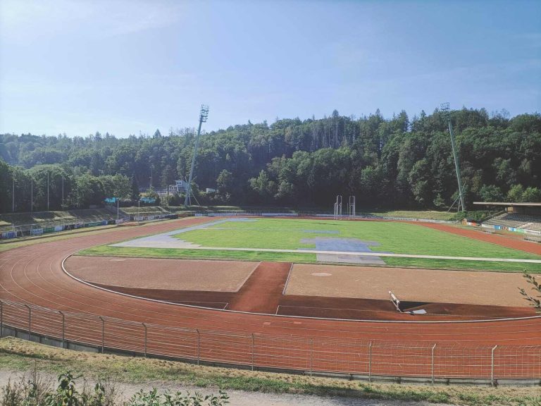 Stadion sportowy z bieżnią, zieloną murawą i otaczającymi go drzewami w tle.