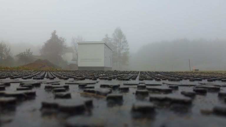 Widok na gładką nawierzchnię drogi, w tle mały budynek w mglistym otoczeniu. Drzewa częściowo zasłonięte mgłą.