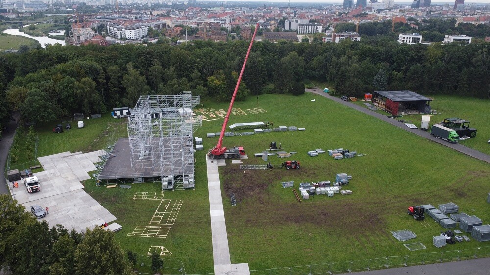 Budowa sceny w parku z rusztowaniem i dźwigiem z długim czerwonym wysięgnikiem.