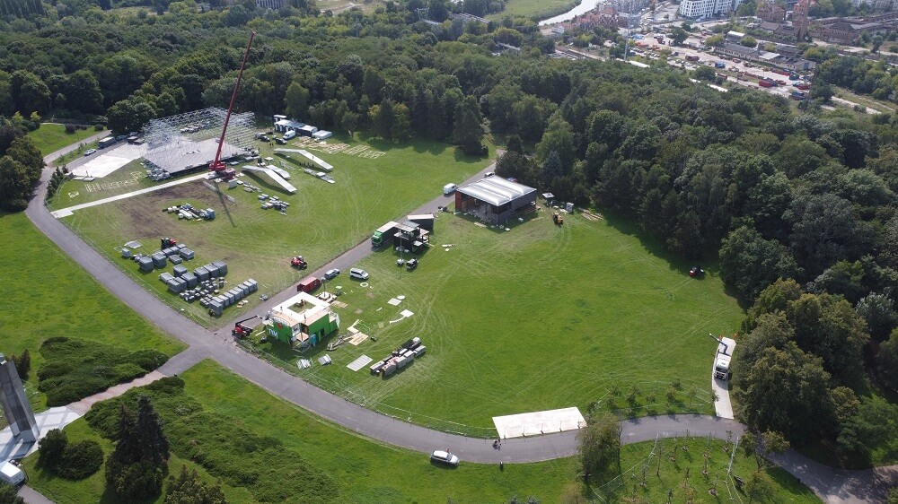 Park z rozstawianą sceną i sprzętem eventowym, dźwig i kontenery.