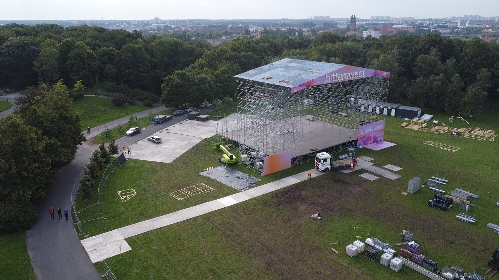 Duża scena z rusztowaniem i banerem bittersweet montowana w parku, ekipa i sprzęt.