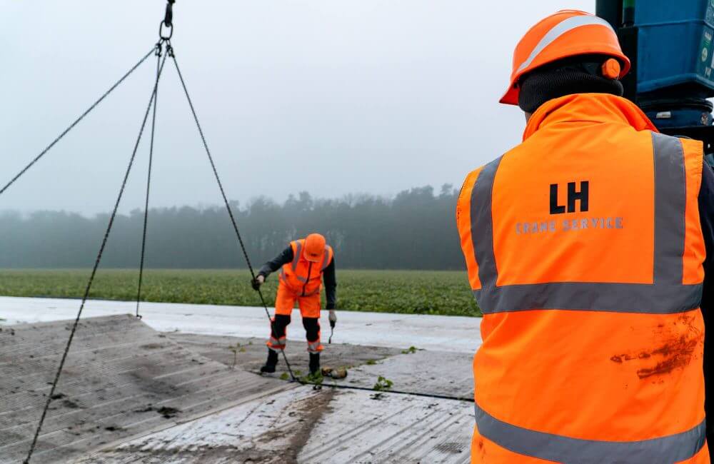Dwaj pracownicy w pomarańczowych kurtkach roboczych na placu budowy, jeden z nich przymocowuje linę do materiału.