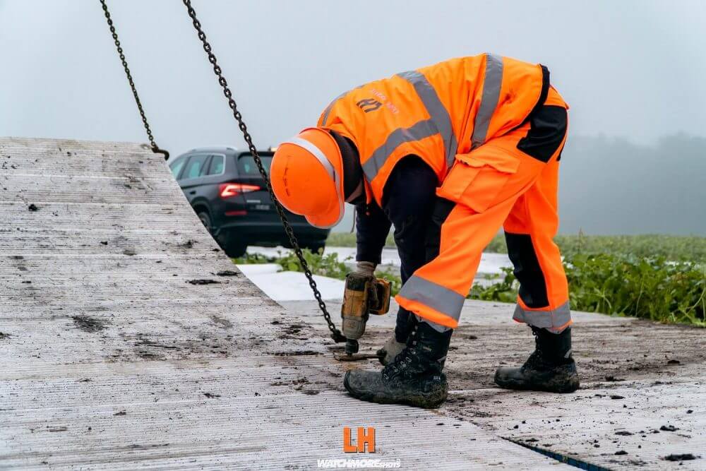 Robotnik w jaskrawej odzieży ochronnej używa wiertarki na placu budowy w mglistym otoczeniu. W tle widać samochód.