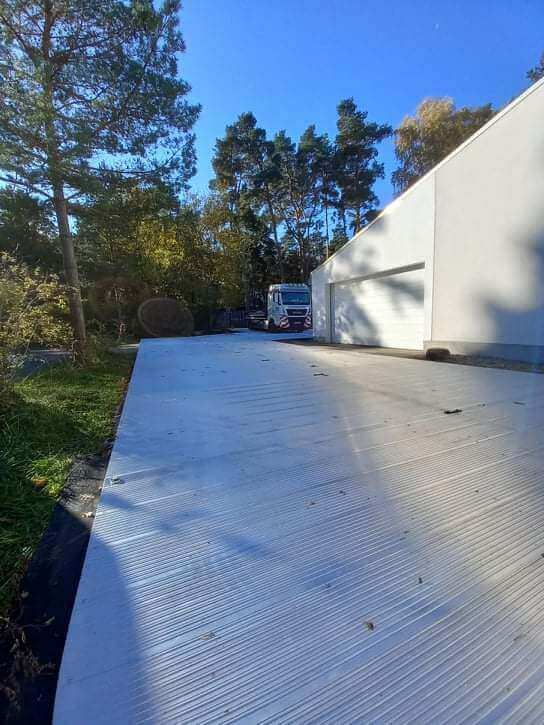 Droga pokryta białymi panelami, z drzewami i budynkiem w tle, lekko oświetlona słońcem.