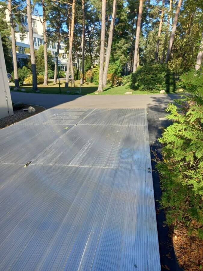 Widok na podjazd z metalową nawierzchnią, otoczony zielonymi drzewami i trawnikiem.