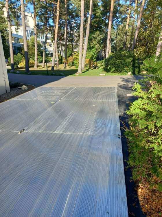 Ścieżka z metalowym pokryciem wśród drzew, z widocznymi budynkami w tle i zieloną roślinnością.