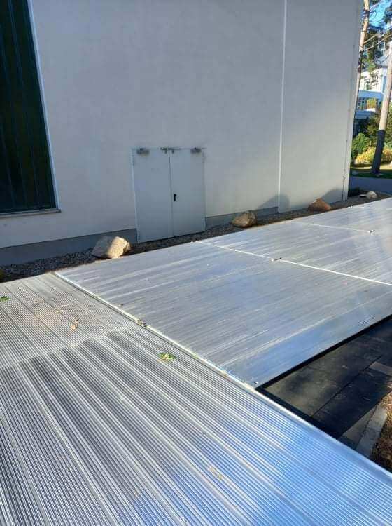 Szklarniowa konstrukcja z przezroczystymi panelami, umieszczona przy budynku, otoczona kamieniami.