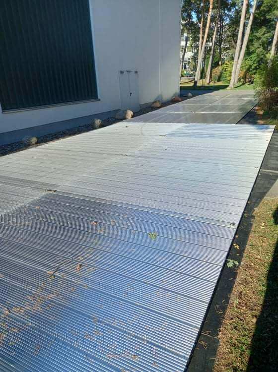 Widok na metalową, żebrowaną nawierzchnię ścieżki w otoczeniu budynku i drzew.