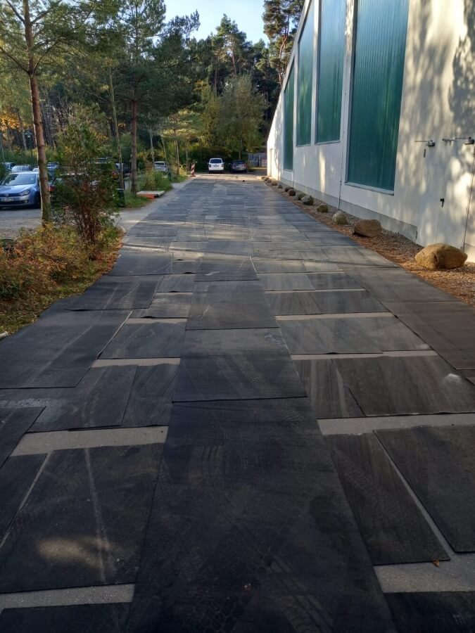 Szersza ścieżka na świeżym powietrzu, wyłożona płytami, z drzewami po bokach i samochodami w tle.