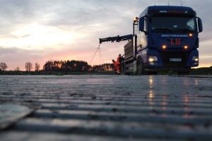 Ciężarówka na drodze z załadunkiem, pracownicy w pomarańczowych ubraniach, zachód słońca w tle.