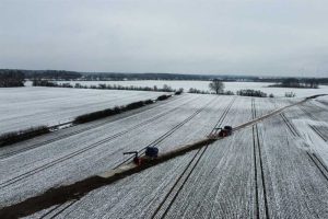 Widok na zimowe pole pokryte śniegiem, z dwiema maszynami na drodze w centralnej części obrazu.