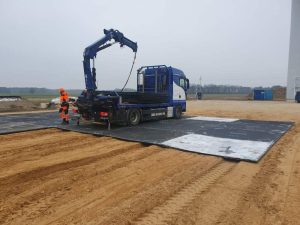Ciężarówka na placu budowy, z podnośnikiem i pracownikiem w odzieży ochronnej. W tle piaszczysta nawierzchnia.