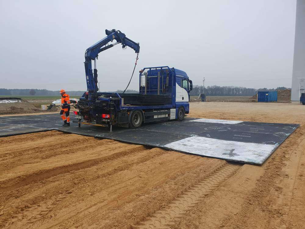 Ciężarówka na placu budowy, z podnośnikiem i pracownikiem w odzieży ochronnej. W tle piaszczysta nawierzchnia.