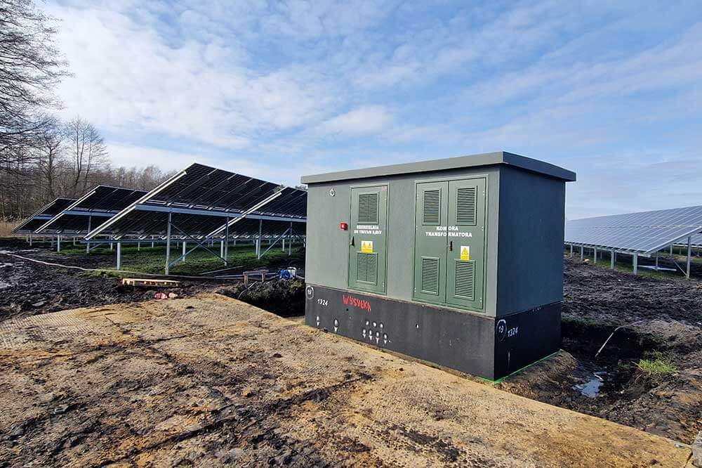 Zespół paneli słonecznych na tle nieba oraz stacja transformacyjna na działce.