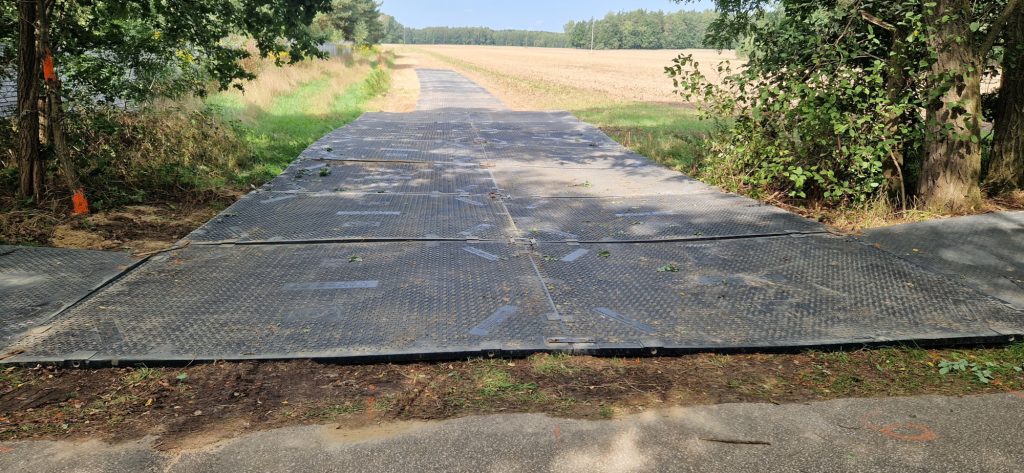 Droga wyłożona czarnymi płytami, otoczona drzewami i polami. W tle widoczny krajobraz wiejski.