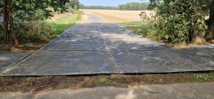 Droga wyłożona czarnymi płytami, otoczona drzewami i polami. W tle widoczny krajobraz wiejski.