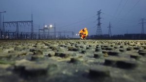 Pracownik w pomarańczowym odblaskowym stroju na zniszczonej powierzchni, w tle linie energetyczne i zmierzch.