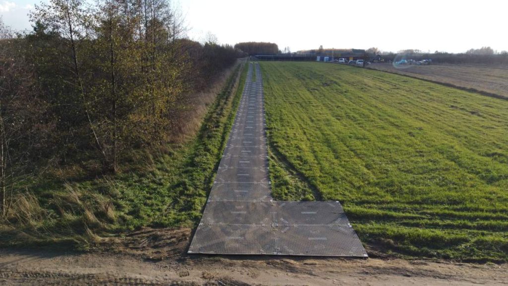 Ścieżka z metalowych płyt prowadząca przez pole, otoczona drzewami i zieloną trawą. W tle widoczne budynki.