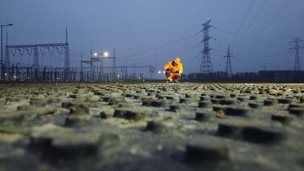 Pracownik w pomarańczowym odblaskowym stroju na zniszczonej powierzchni, w tle linie energetyczne i zmierzch.