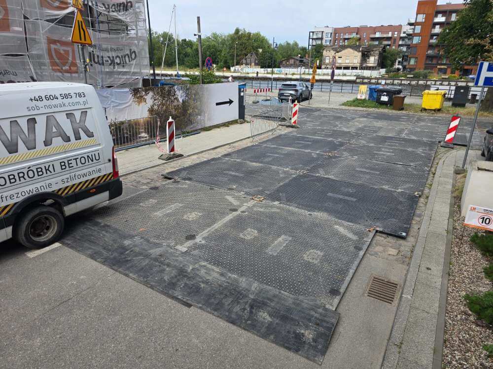 Placyk budowlany z rozłożoną nawierzchnią z płyt, zaparkowanym vanem i oznakowaniem robót drogowych.