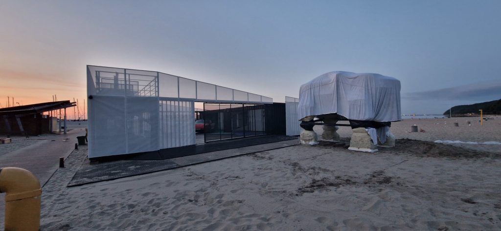 Nowoczesna konstrukcja na plaży, częściowo przykryta materiałem, otoczona piaskiem i w tle zachód słońca.
