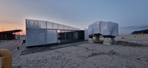 Nowoczesna konstrukcja na plaży, częściowo przykryta materiałem, otoczona piaskiem i w tle zachód słońca.