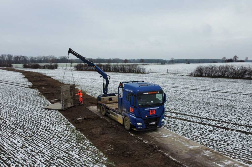 Ciężarówka z dźwigiem podnosi betonowy element na polu w zimowej scenerii.
