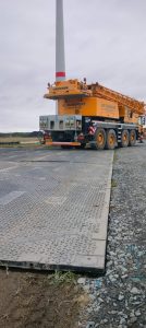 Żółty dźwig na dużej płycie transportowej, obok widoczny wieżowiec wiatrowy i pole. Chmurne niebo w tle.